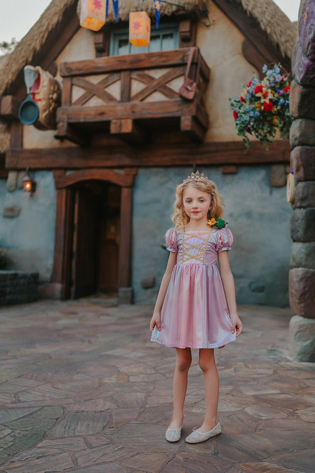 A young girls is wearing a metallic Rapunzel Twirl dress. The pink and purple knee length dress has a criss cross gold ribbon across the bodice. The ballroom puff sleeves add elegance.