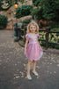 A young girls is wearing a metallic Rapunzel Twirl dress. The pink and purple knee length dress has a criss cross gold ribbon across the bodice. The ballroom puff sleeves add elegance.