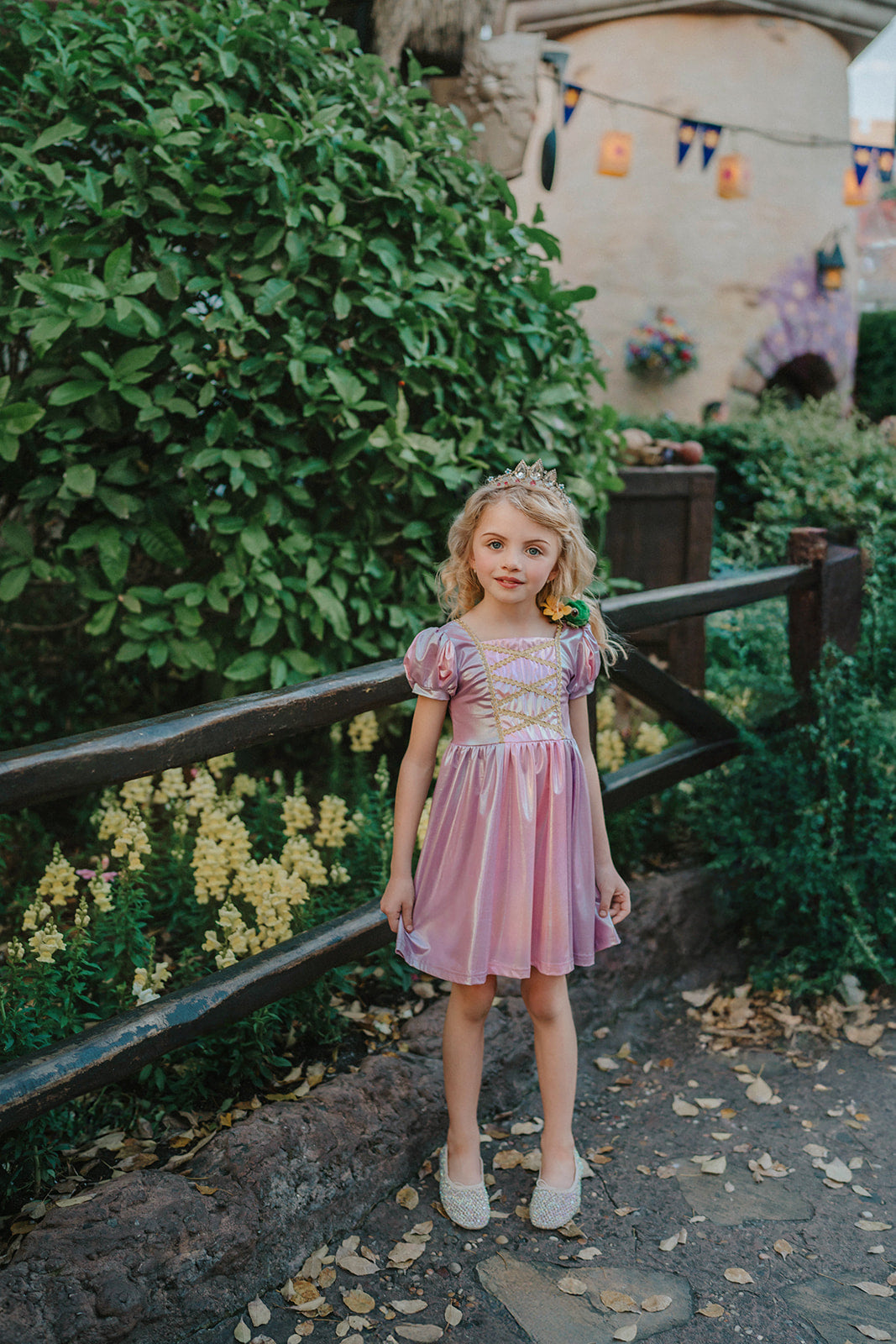 A young girls is wearing a metallic Rapunzel Twirl dress. The pink and purple knee length dress has a criss cross gold ribbon across the bodice. The ballroom puff sleeves add elegance.