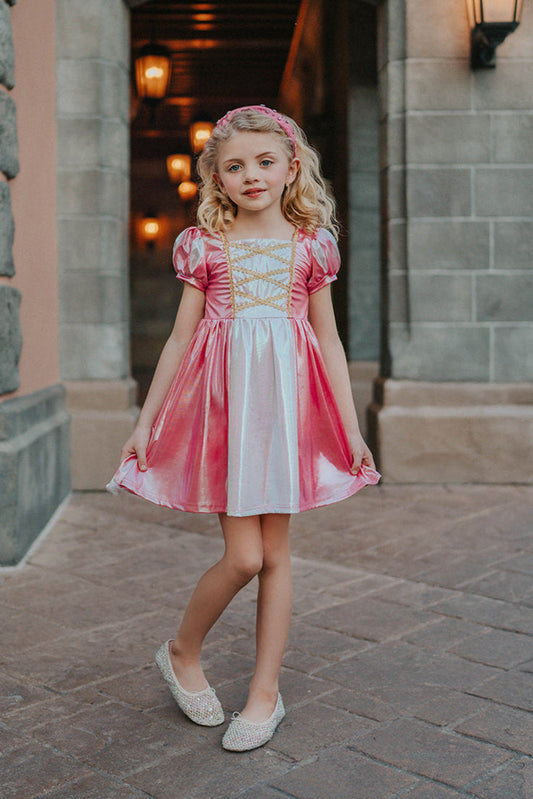 A young girls is wearing a metallic Sleeping Beauty Twirl dress. The pink  and white knee length dress has a criss cross gold ribbon across the bodice. The ballroom puff sleeves add elegance.