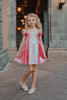 A young girls is wearing a metallic Sleeping Beauty Twirl dress. The pink  and white knee length dress has a criss cross gold ribbon across the bodice. The ballroom puff sleeves add elegance.