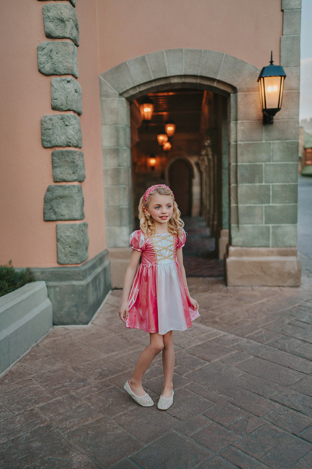 A young girls is wearing a metallic Sleeping Beauty Twirl dress. The pink  and white knee length dress has a criss cross gold ribbon across the bodice. The ballroom puff sleeves add elegance.