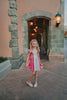 A young girls is wearing a metallic Sleeping Beauty Twirl dress. The pink  and white knee length dress has a criss cross gold ribbon across the bodice. The ballroom puff sleeves add elegance.