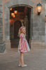 A young girls is wearing a metallic Sleeping Beauty Twirl dress. The pink  and white knee length dress has a criss cross gold ribbon across the bodice. The ballroom puff sleeves add elegance.