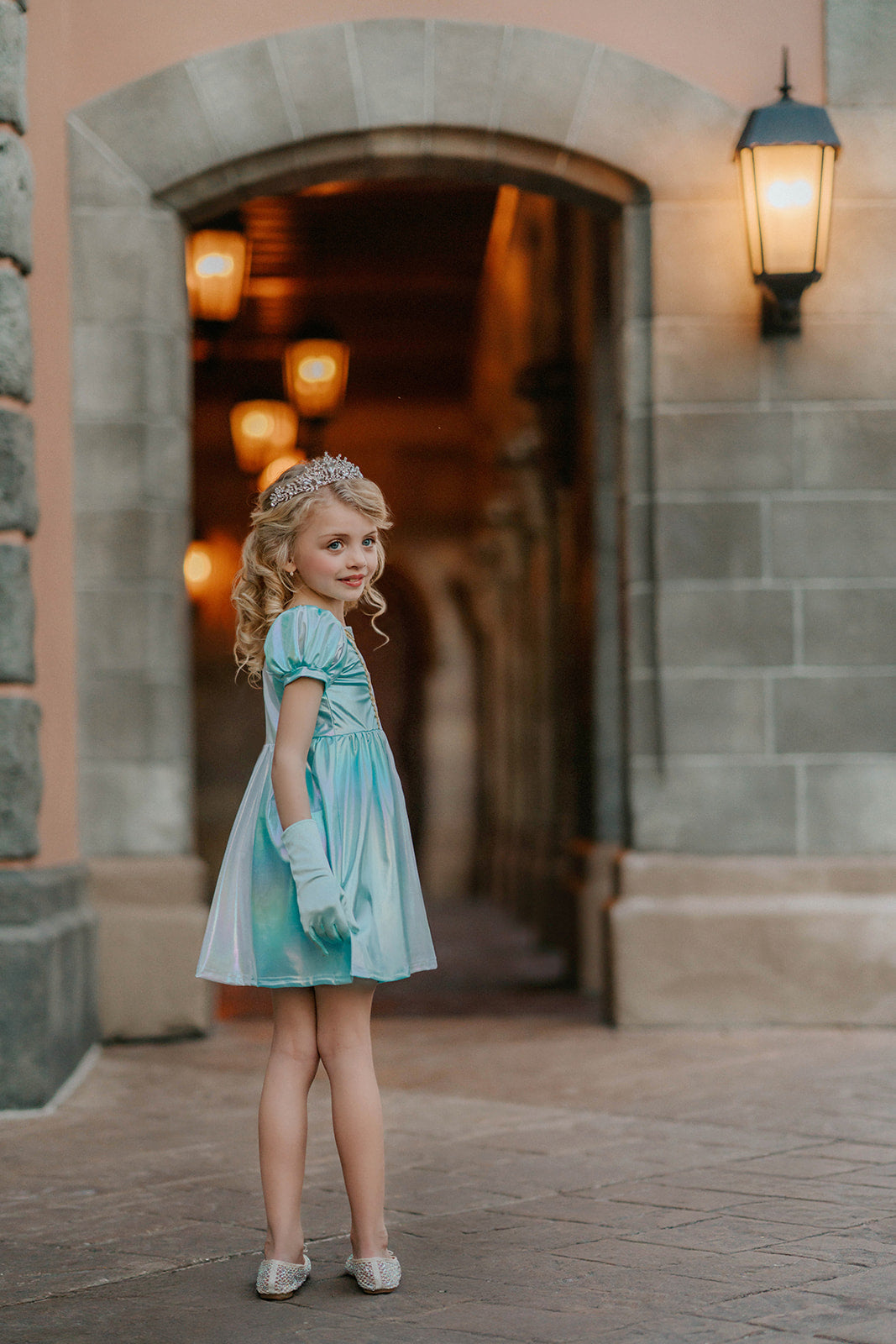 A young girls is wearing a metallic Cinderella Twirl dress. The light blue and white knee length dress has a criss cross gold ribbon across the bodice. The ballroom puff sleeves add elegance.