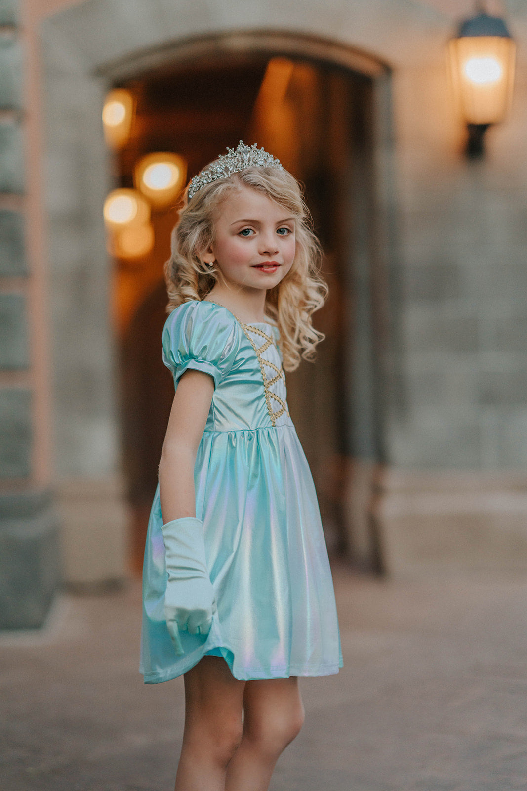 A young girls is wearing a metallic Cinderella Twirl dress. The light blue and white knee length dress has a criss cross gold ribbon across the bodice. The ballroom puff sleeves add elegance.