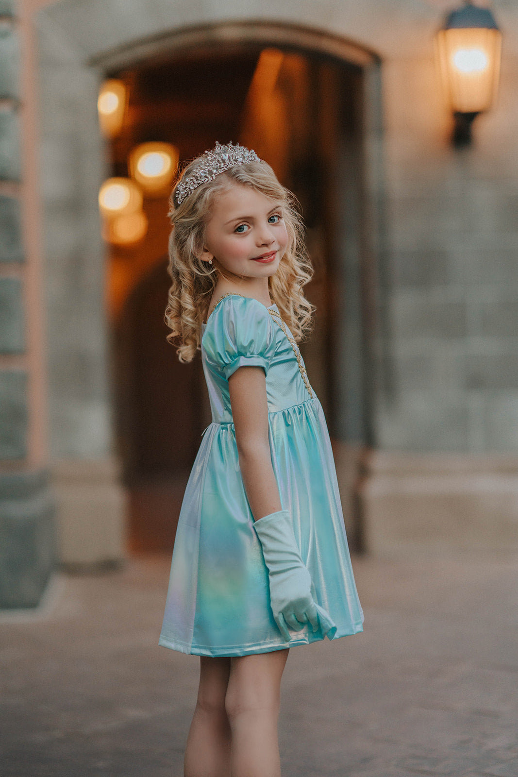 A young girls is wearing a metallic Cinderella Twirl dress. The light blue and white knee length dress has a criss cross gold ribbon across the bodice. The ballroom puff sleeves add elegance.