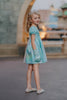 A young girls is wearing a metallic Cinderella Twirl dress. The light blue and white knee length dress has a criss cross gold ribbon across the bodice. The ballroom puff sleeves add elegance.