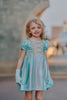 A young girls is wearing a metallic Cinderella Twirl dress. The light blue and white knee length dress has a criss cross gold ribbon across the bodice. The ballroom puff sleeves add elegance.