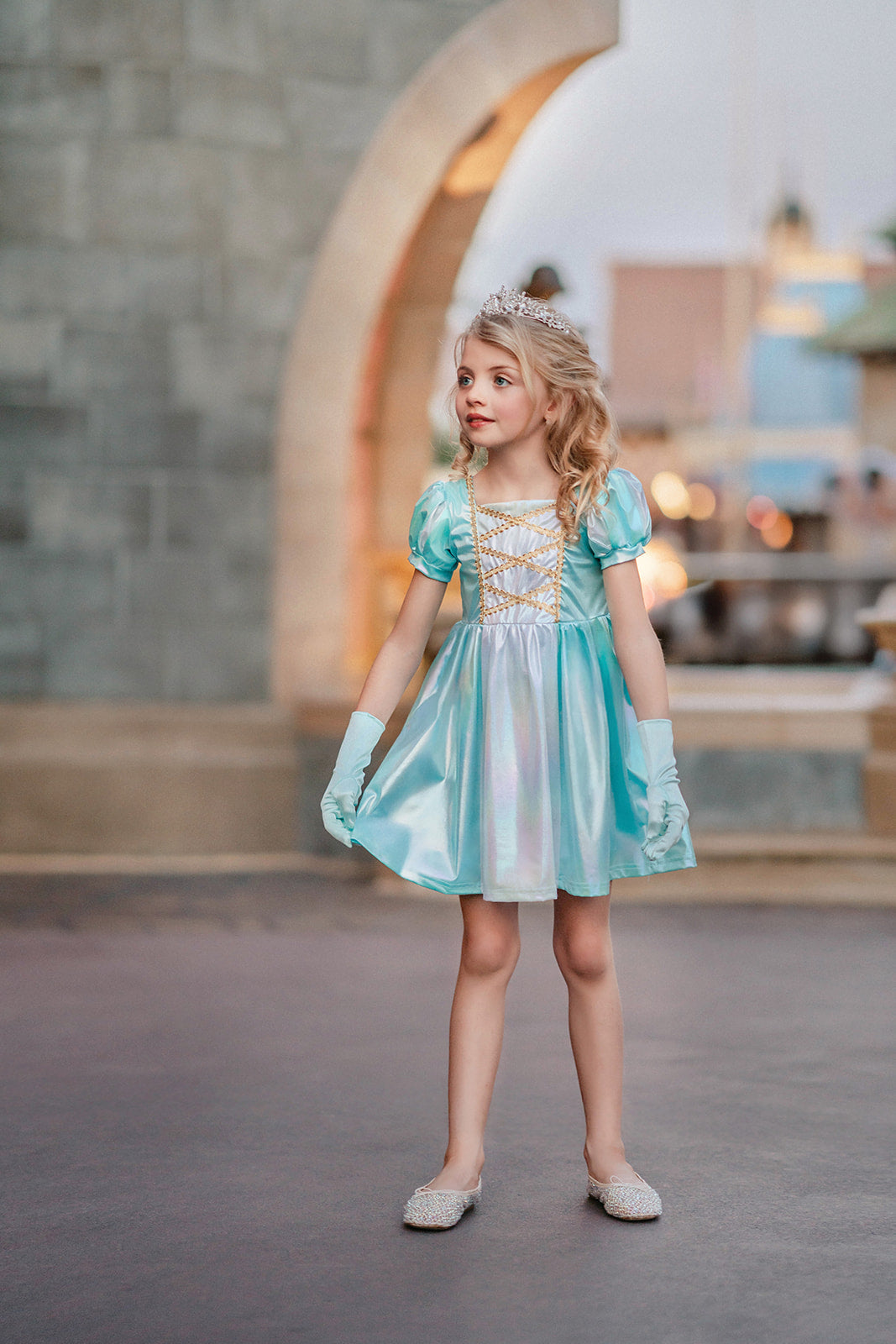 A young girls is wearing a metallic Cinderella Twirl dress. The light blue and white knee length dress has a criss cross gold ribbon across the bodice. The ballroom puff sleeves add elegance.