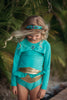 A young girl is wearing a bight teal and gold rash guard swim set.  The top is long sleeved and at the hem as a gold band and teal ruffles. There is a teal blue and gold jewel at the neckline.  The bottom is a modest cut with gold waist and teal. 