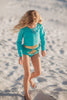 A young girl is wearing a bight teal and gold rash guard swim set.  The top is long sleeved and at the hem as a gold band and teal ruffles. There is a teal blue and gold jewel at the neckline.  The bottom is a modest cut with gold waist and teal. 