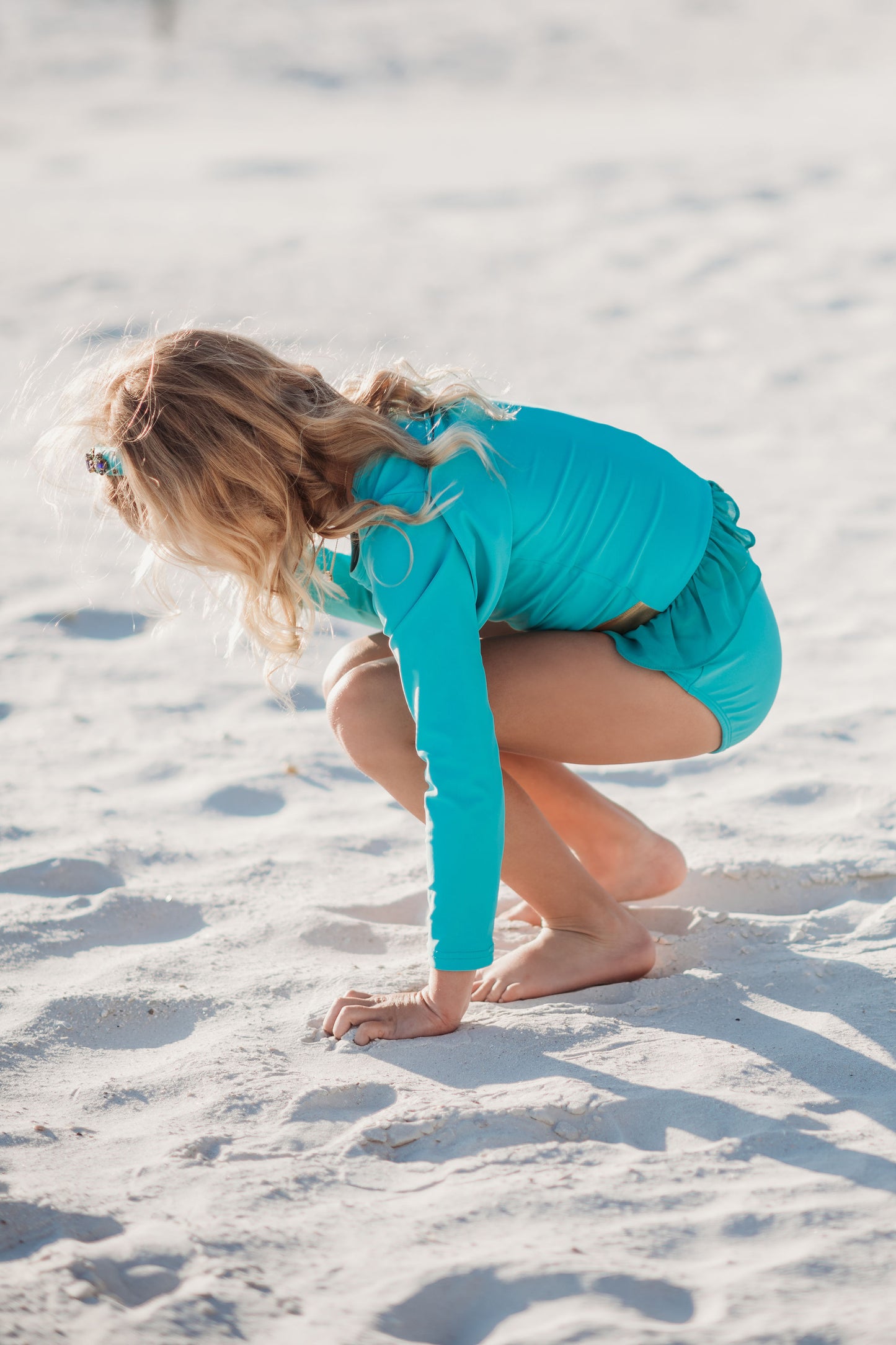 A young girl is wearing a bight teal and gold rash guard swim set.  The top is long sleeved and at the hem as a gold band and teal ruffles. There is a teal blue and gold jewel at the neckline.  The bottom is a modest cut with gold waist and teal. 