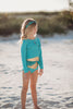 A young girl is wearing a bight teal and gold rash guard swim set.  The top is long sleeved and at the hem as a gold band and teal ruffles. There is a teal blue and gold jewel at the neckline.  The bottom is a modest cut with gold waist and teal. 