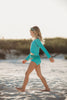 A young girl is wearing a bight teal and gold rash guard swim set.  The top is long sleeved and at the hem as a gold band and teal ruffles. There is a teal blue and gold jewel at the neckline.  The bottom is a modest cut with gold waist and teal. 