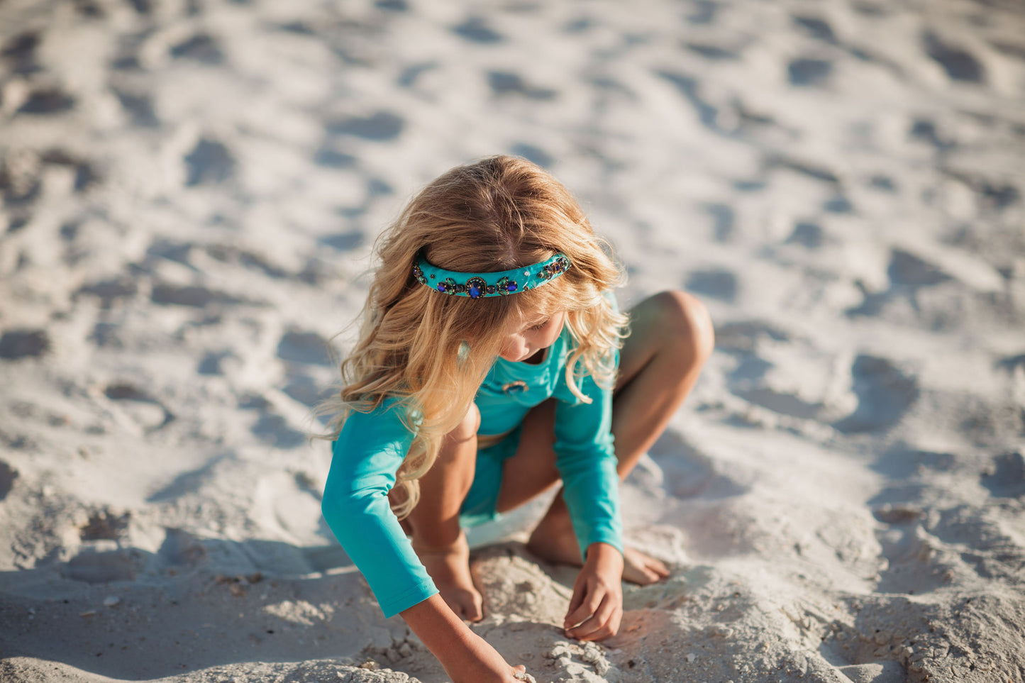 A young girl is wearing a bight teal and gold rash guard swim set.  The top is long sleeved and at the hem as a gold band and teal ruffles. There is a teal blue and gold jewel at the neckline.  The bottom is a modest cut with gold waist and teal. 