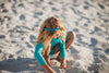 A young girl is wearing a bight teal and gold rash guard swim set.  The top is long sleeved and at the hem as a gold band and teal ruffles. There is a teal blue and gold jewel at the neckline.  The bottom is a modest cut with gold waist and teal. 