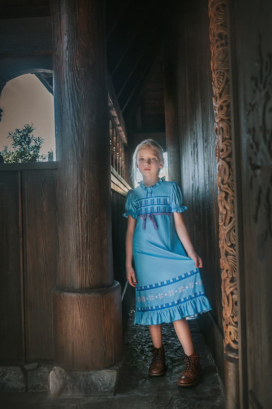 A girl stands in our buttery soft Ice Queen Gown.  A blue nightgown with Scandinavian floral print. Short puff sleeves and a ruffle at the hem.  