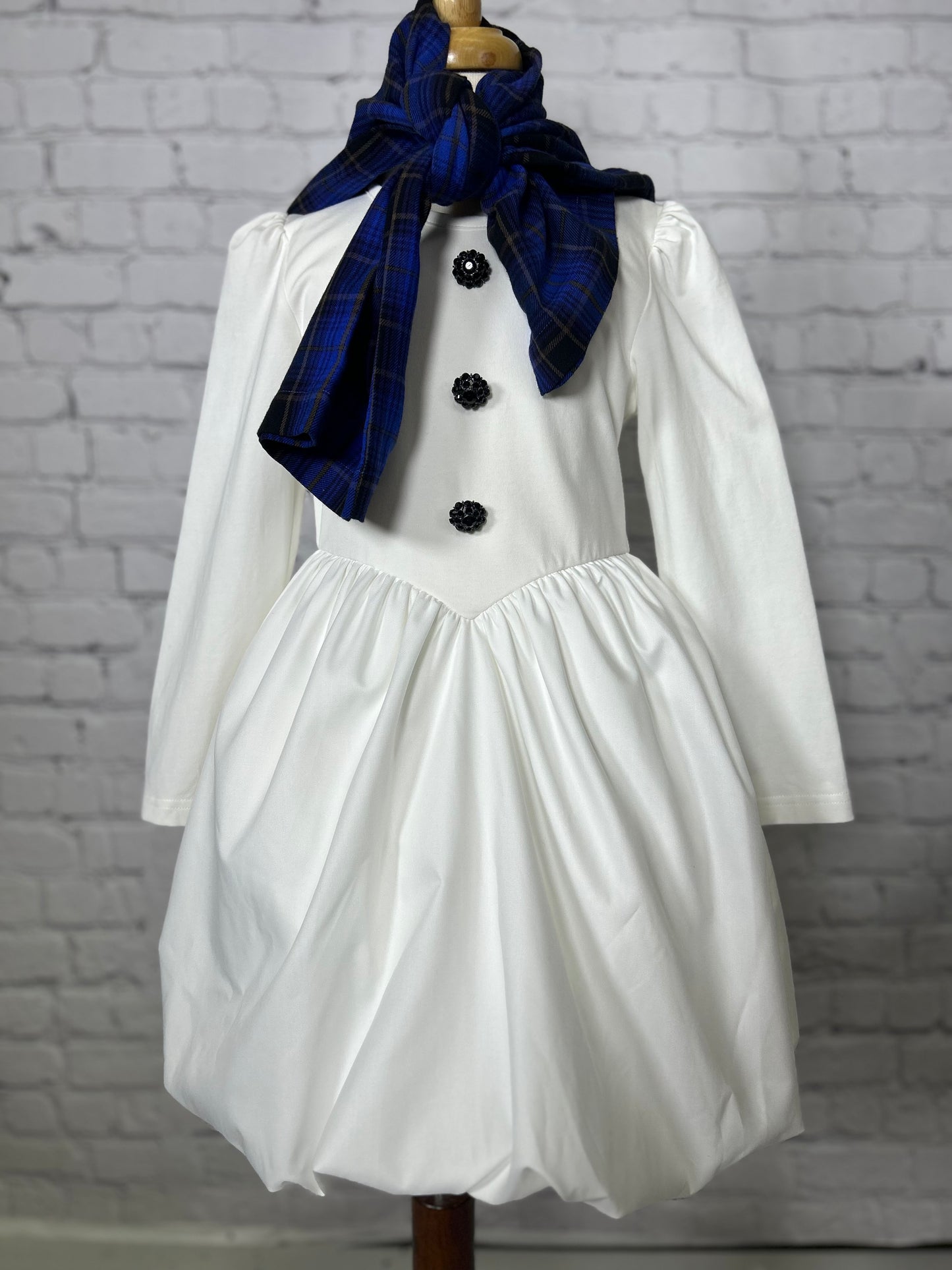 Girl wearing white holiday dress with bubble hem, black tights, and a plaid scarf, styled as a snowman outfit for Christmas photos, winter events, or holiday parties.

Child in snowman-inspired winter dress with black leggings, puff hem skirt, and blue plaid scarf, perfect for Christmas card photos and seasonal festivities.