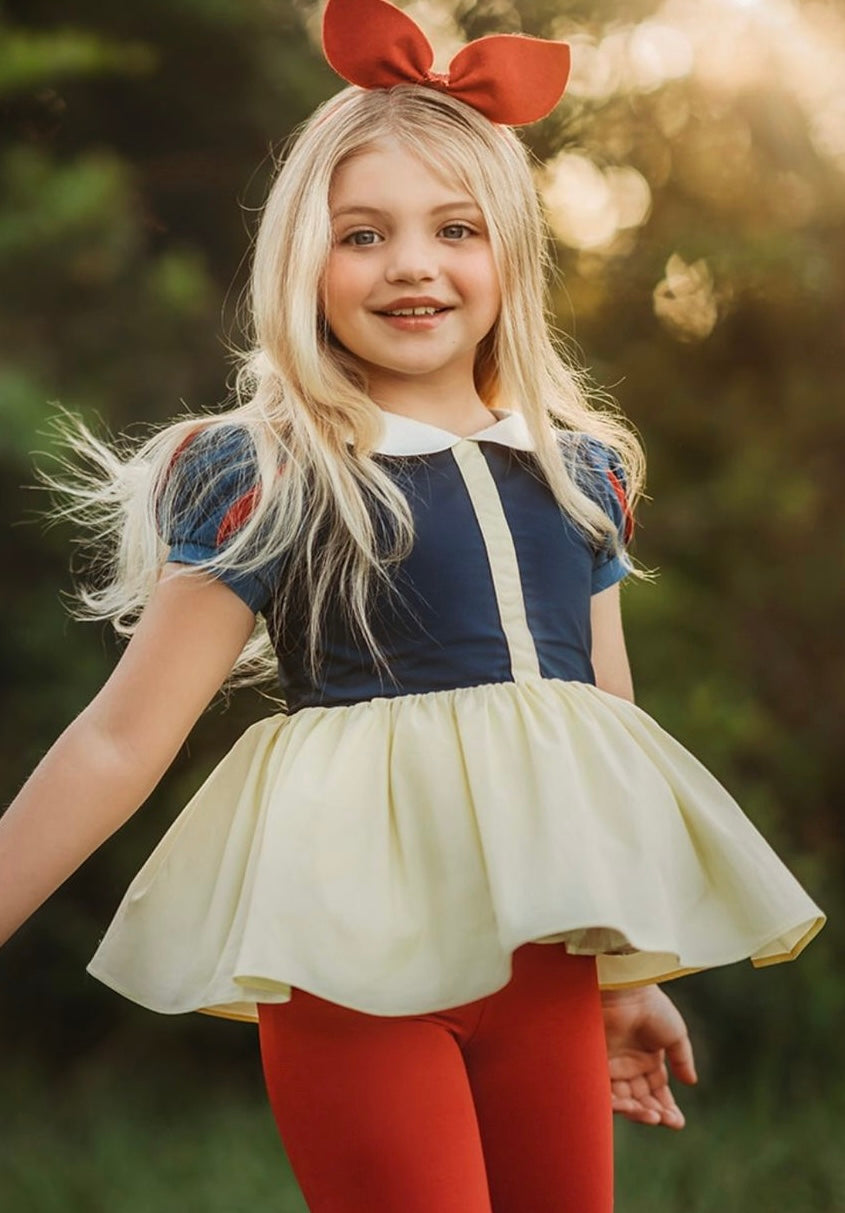 A young girl is wearing the Snow White Peplum top.  The top has a white peter pan collar and red and blue puff sleeves.  The navy blue bodice has a yellow accent and leads into a light yellow peplum skirt.  There is tulle under with a cotton liner to add fullness. 