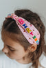 The light pink satin headband has "Happy Birthday" spelled out in colorful beading.  Each letter is a bight color of pink, blue, yellow, green or orange. There are multicolor rhinestones as well. The center has a small tied knot.  This hair accessory is designed for all day comfort. 