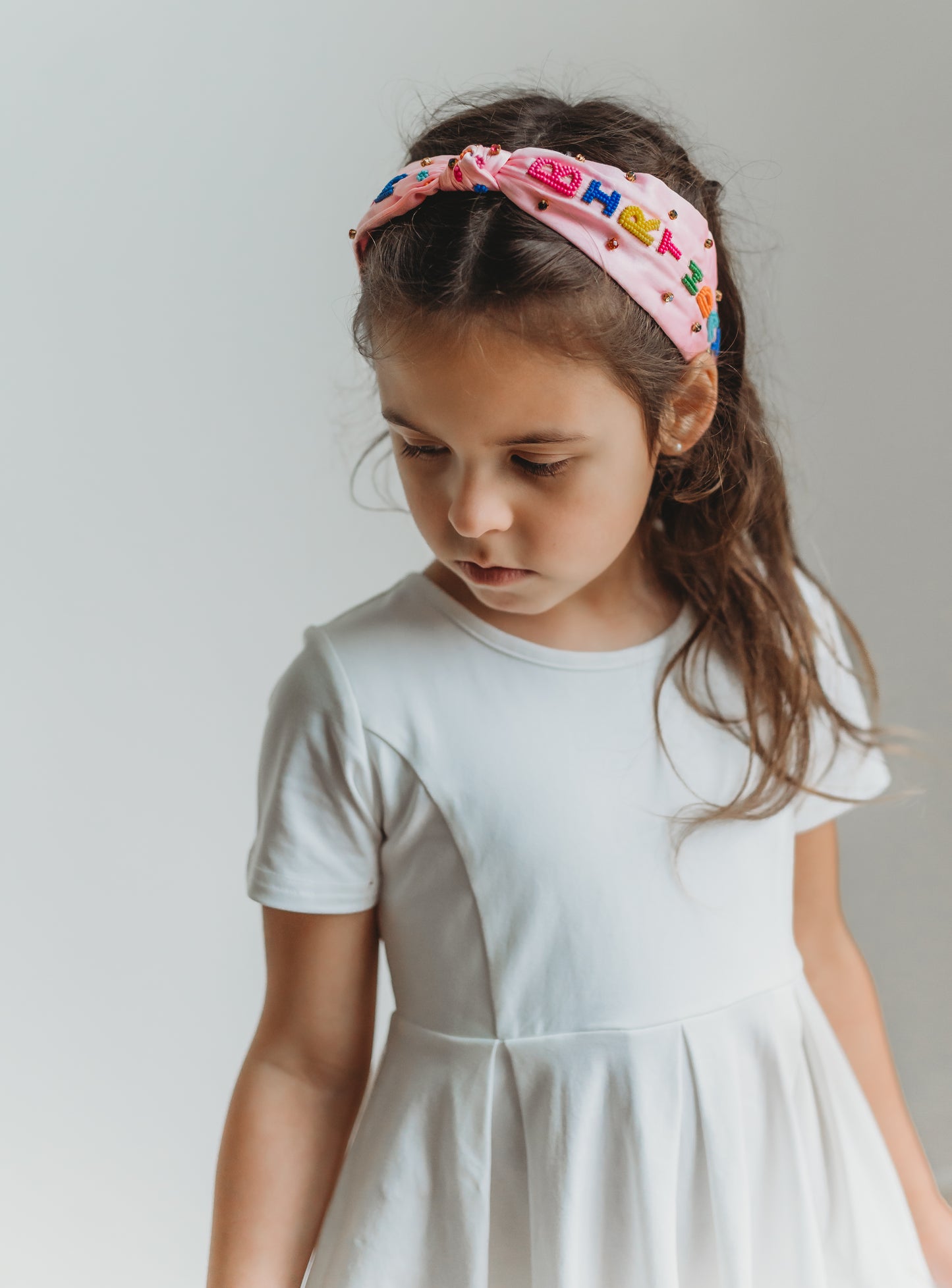 The light pink satin headband has "Happy Birthday" spelled out in colorful beading.  Each letter is a bight color of pink, blue, yellow, green or orange. There are multicolor rhinestones as well. The center has a small tied knot.  This hair accessory is designed for all day comfort. 