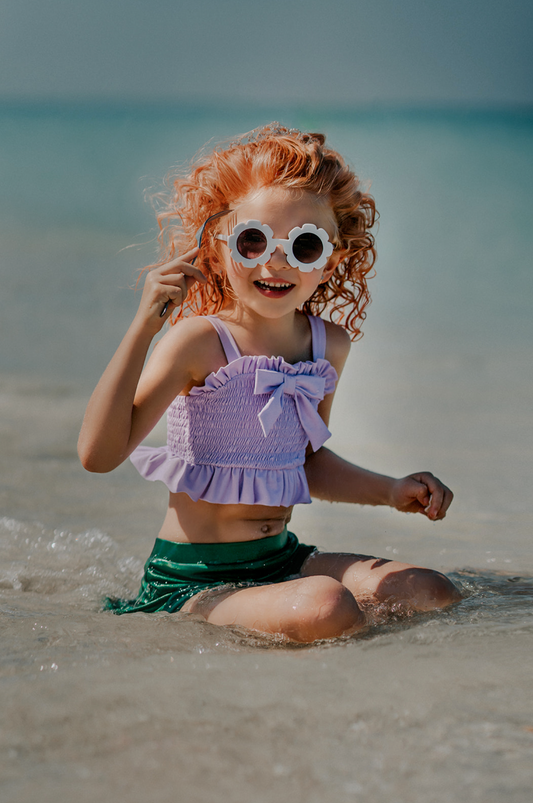 A young girl playing in the water is wearing the Mermaid Tail 2pc Swim set.  The top is light purple with ruched bodice and ruffles.  There is a bow in the center.  The metallic green bottom is cut to resemble a mermaid and is longer on the sides.  