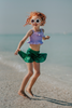 A young girl playing in the water is wearing the Mermaid Tail 2pc Swim set.  The top is light purple with ruched bodice and ruffles.  There is a bow in the center.  The metallic green bottom is cut to resemble a mermaid and is longer on the sides.  