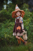 A young girl wears a tan long sleeve cotton Scarecrow dress.  The dress has a jester triangle collar in grey.  There is an overall brown apron with handkerchief bottom.  There are orange and blue plaid patches on the sleeves and apron. 
