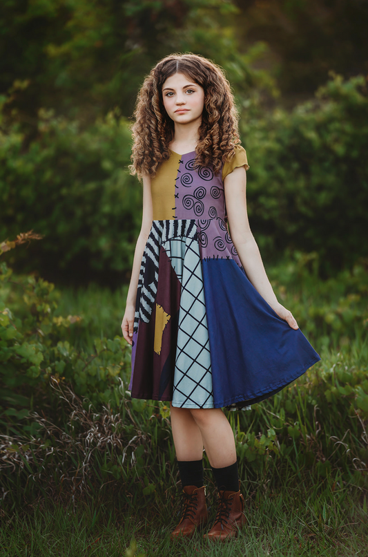 A teen girl is wearing "The Pumpkin King's Friend" dress, its colorful patchwork in purple, black, and mustard yellow made of soft poly-spandex blend fabric. Perfect for spooky season!