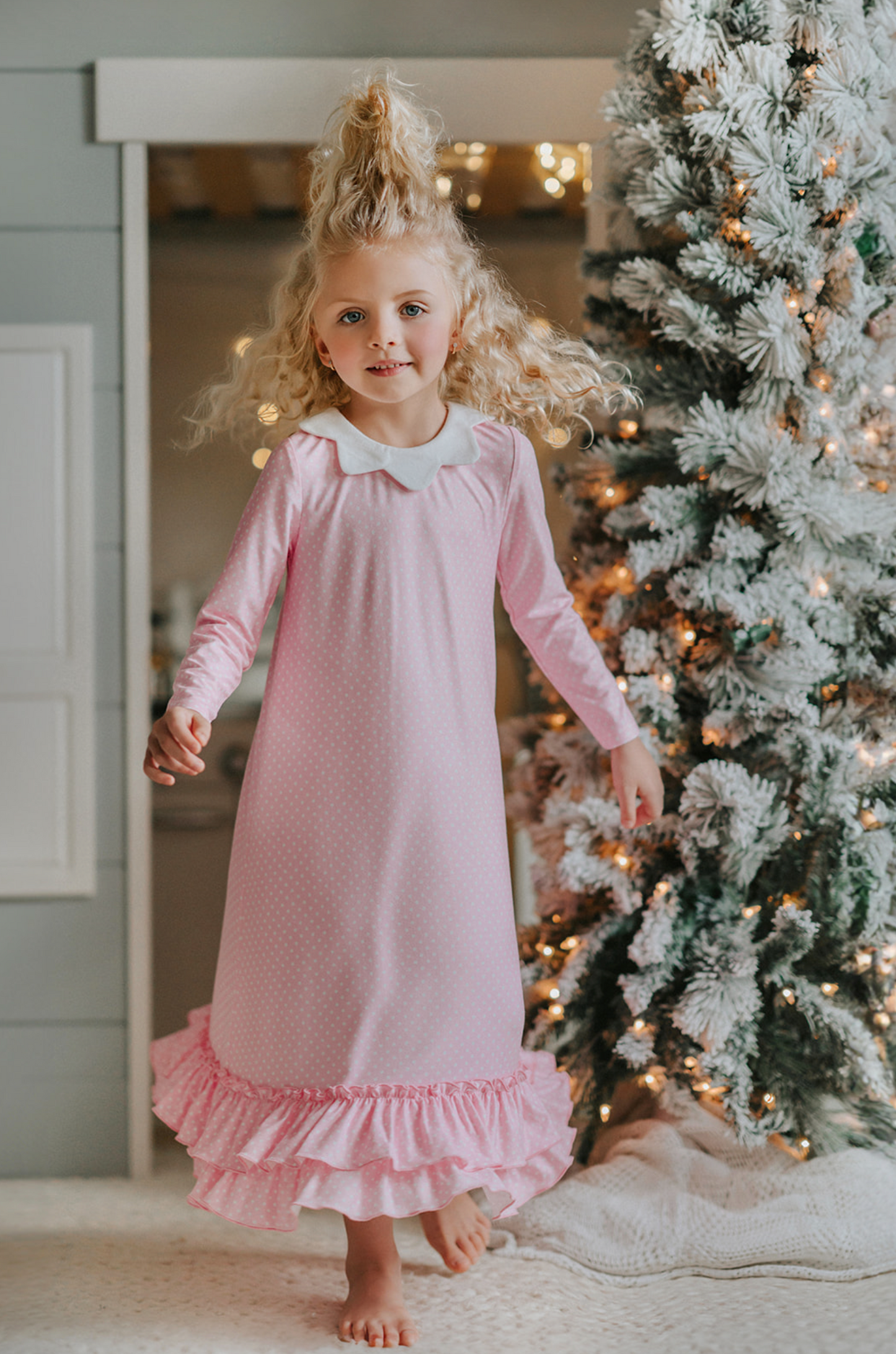 A young girl is wearing the Sleepy Who Gown inspired by Cindy Lou Who.  The buttery soft lounge wear has a white collar.  Light pink with small white polka dots and two adorable ruffles at the bottom. 