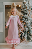 A young girl is wearing the Sleepy Who Gown inspired by Cindy Lou Who.  The buttery soft lounge wear has a white collar.  Light pink with small white polka dots and two adorable ruffles at the bottom. 