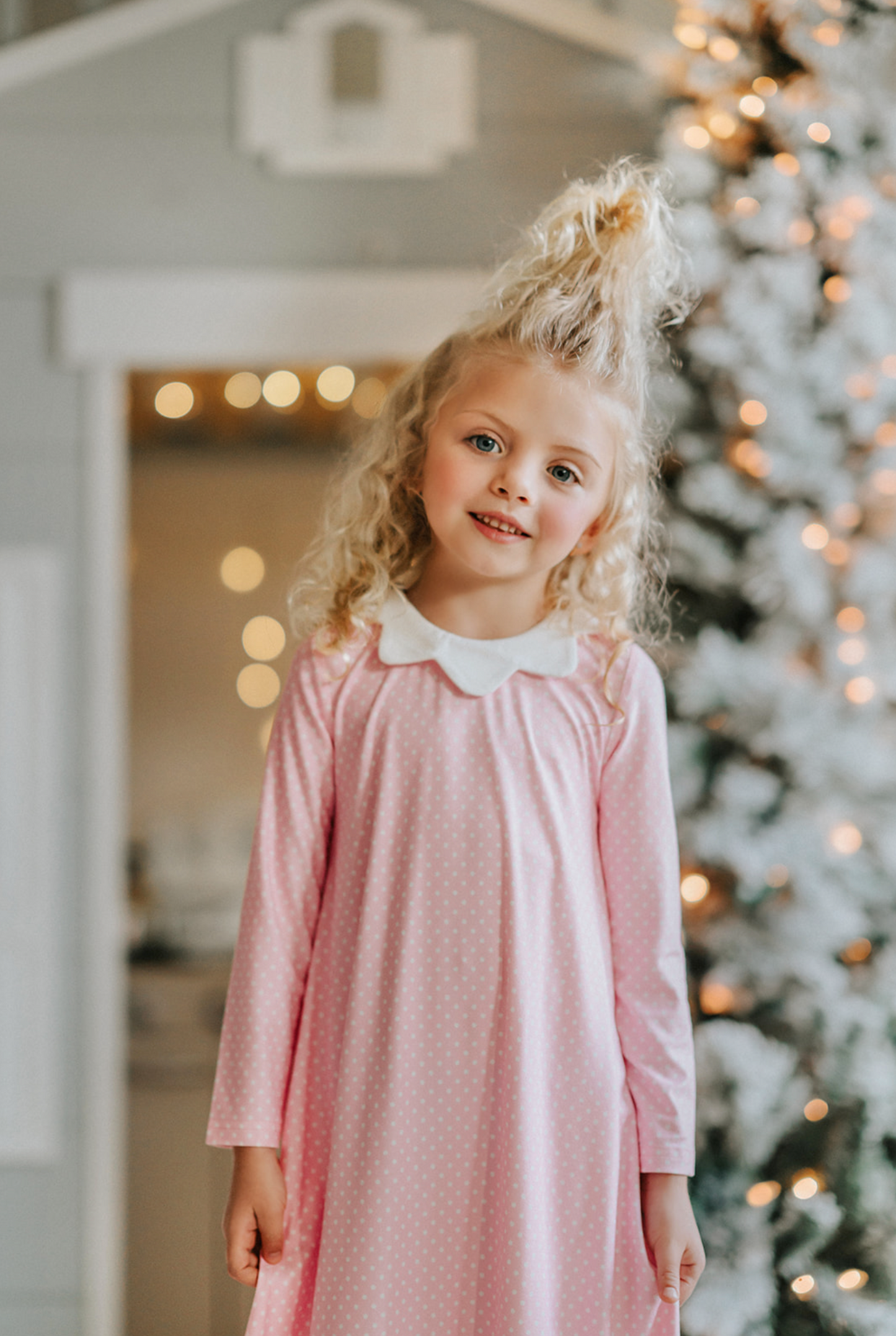 A young girl is wearing the Sleepy Who Gown inspired by Cindy Lou Who.  The buttery soft lounge wear has a white collar.  Light pink with small white polka dots and two adorable ruffles at the bottom. 