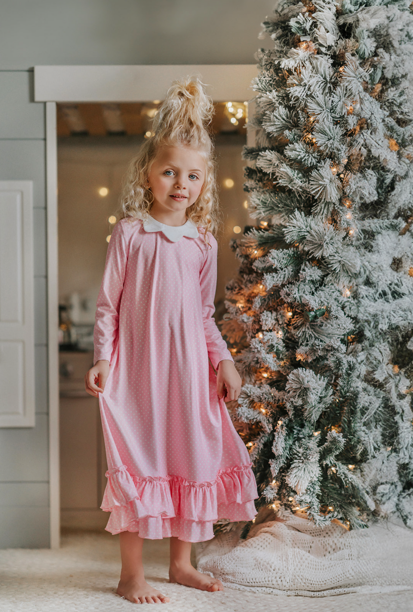 A young girl is wearing the Sleepy Who Gown inspired by Cindy Lou Who.  The buttery soft lounge wear has a white collar.  Light pink with small white polka dots and two adorable ruffles at the bottom. 