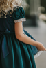 A young girl stands indoors wearing a dark teal green dress with white lace trim, a matching plaid blue and green apron, puff sleeves and a white collar.  The woven cotton is perfect for spooky season.