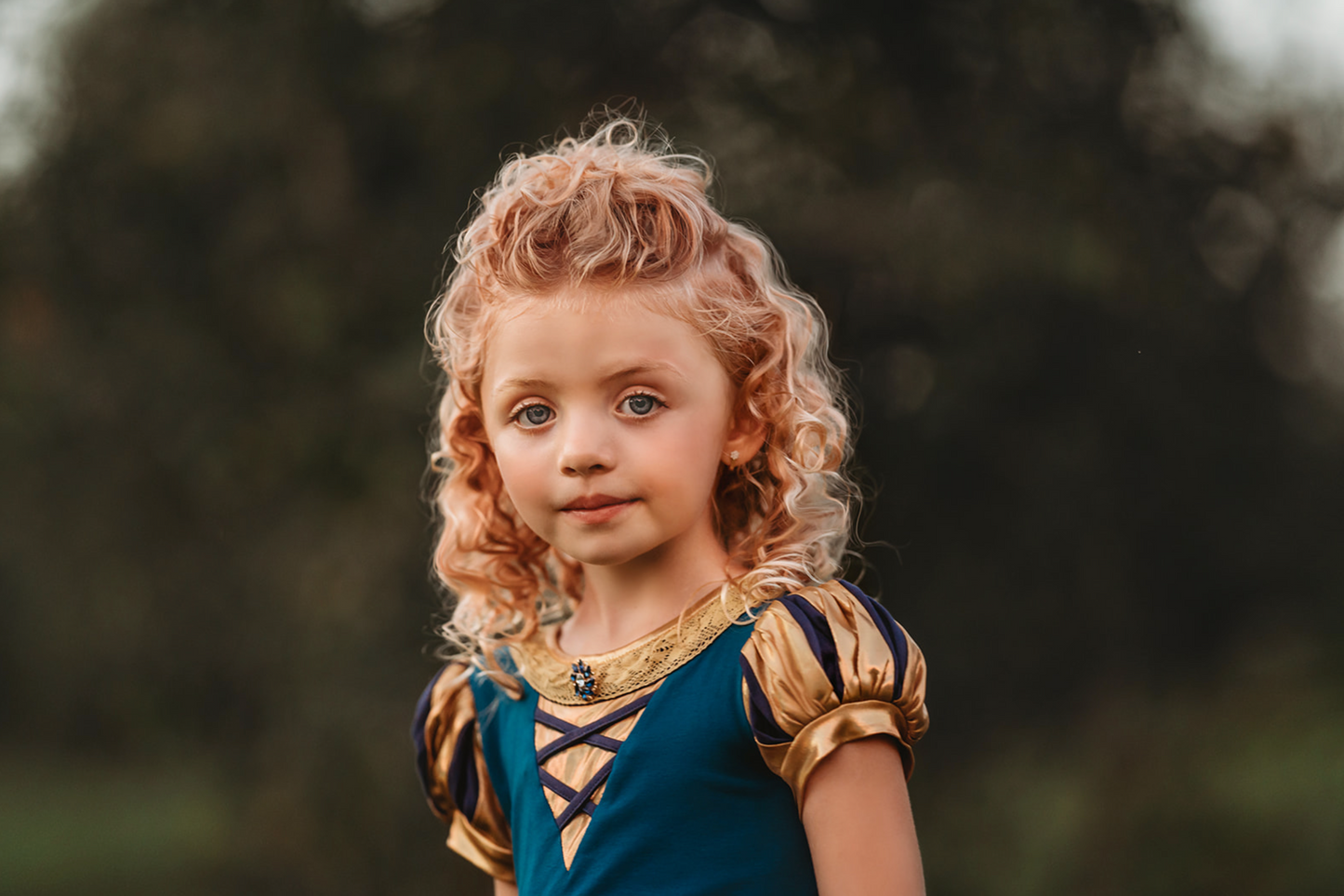 A young girl is wearing a dark teal cotton twirl dress.  The puff sleeves have metallic gold.  Metallic gold lines the neck with a dark teal jewel.  A intricate gold ribbon runs along the hem. 