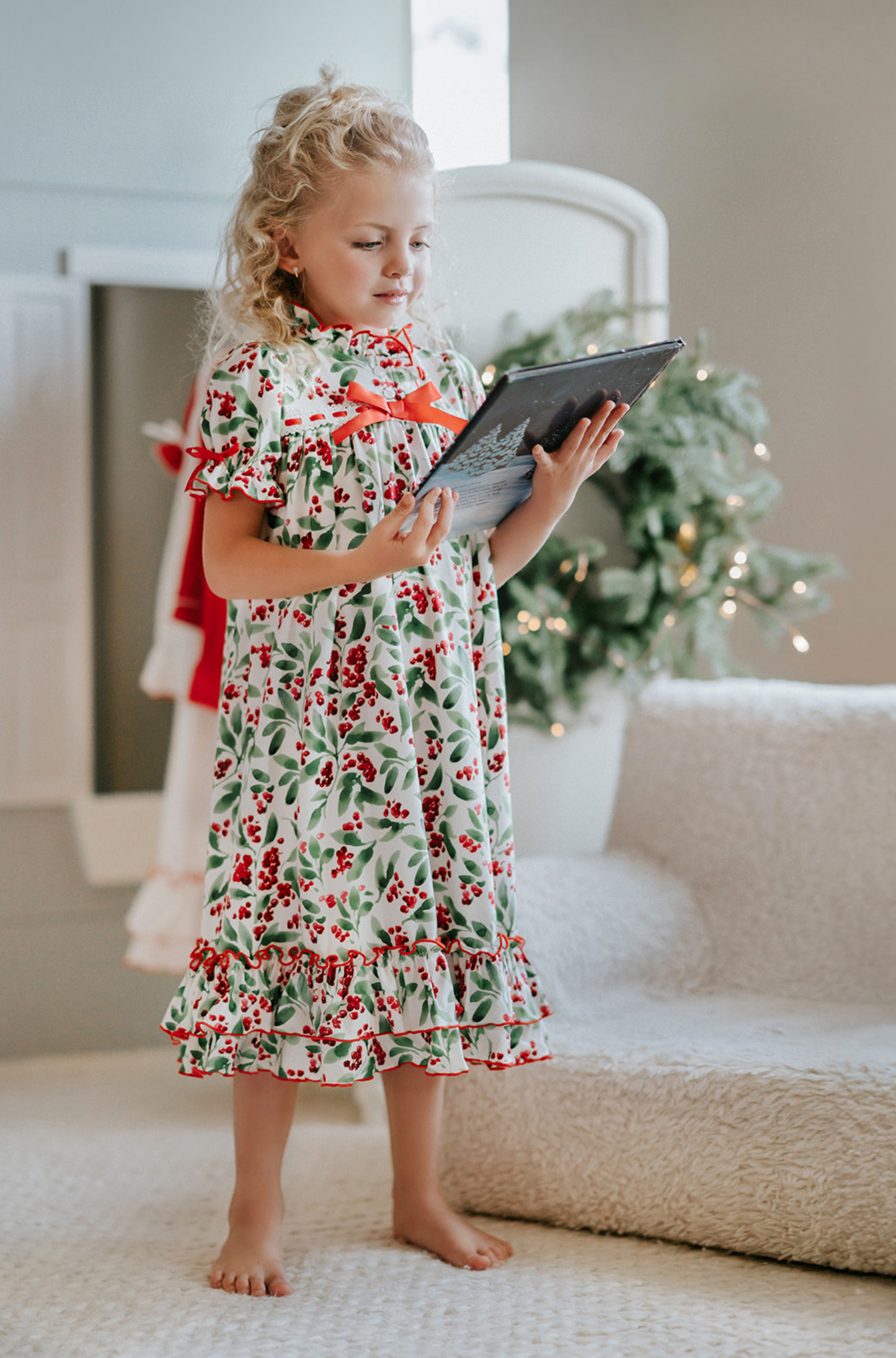 A young girl is wearing The Original Clara Nutcracker Gown in Winterberry, featuring winterberry print and a red bow. The buttery soft fabric is cozy and sensory friendly.  The puff sleeves with ribbon detail and the ruffles at the hem add a traditional classic look. 