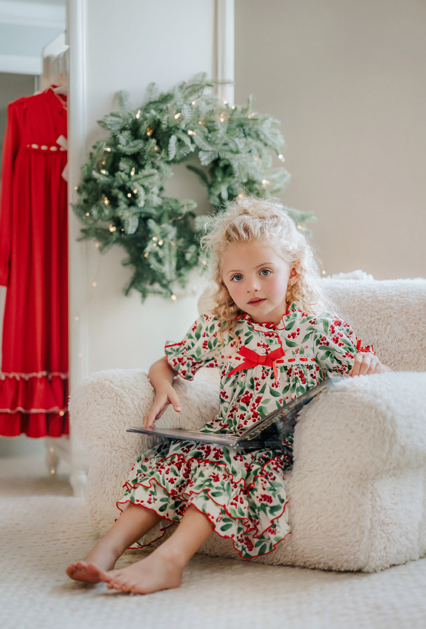 A young child with curly blonde hair stands barefoot on a soft carpet, holding an open book. The child wears *The Original Clara Nutcracker Gown in Winterberry* and reads intently. A decorated Christmas tree is visible in the background, part of our exclusive Christmas Lounge Collection.