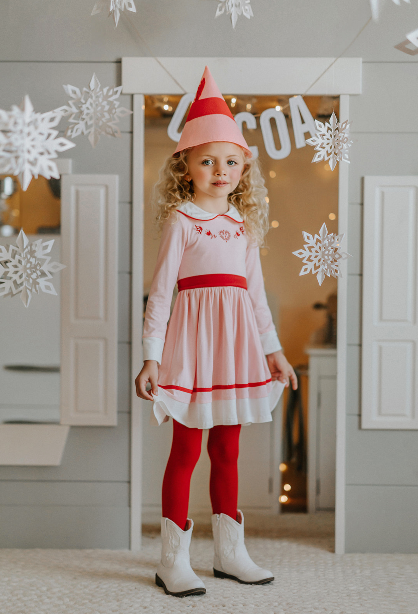 A young girl is wearing a long sleeve cotton dress in a light pink.  The peter pan collar has red trim. The chest has red embroidered accents.  The skirt has a white and red band at the hemline. 