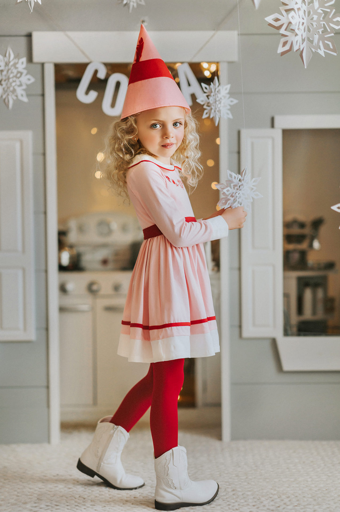 A young girl is wearing a long sleeve cotton dress in a light pink.  The peter pan collar has red trim. The chest has red embroidered accents.  The skirt has a white and red band at the hemline. 