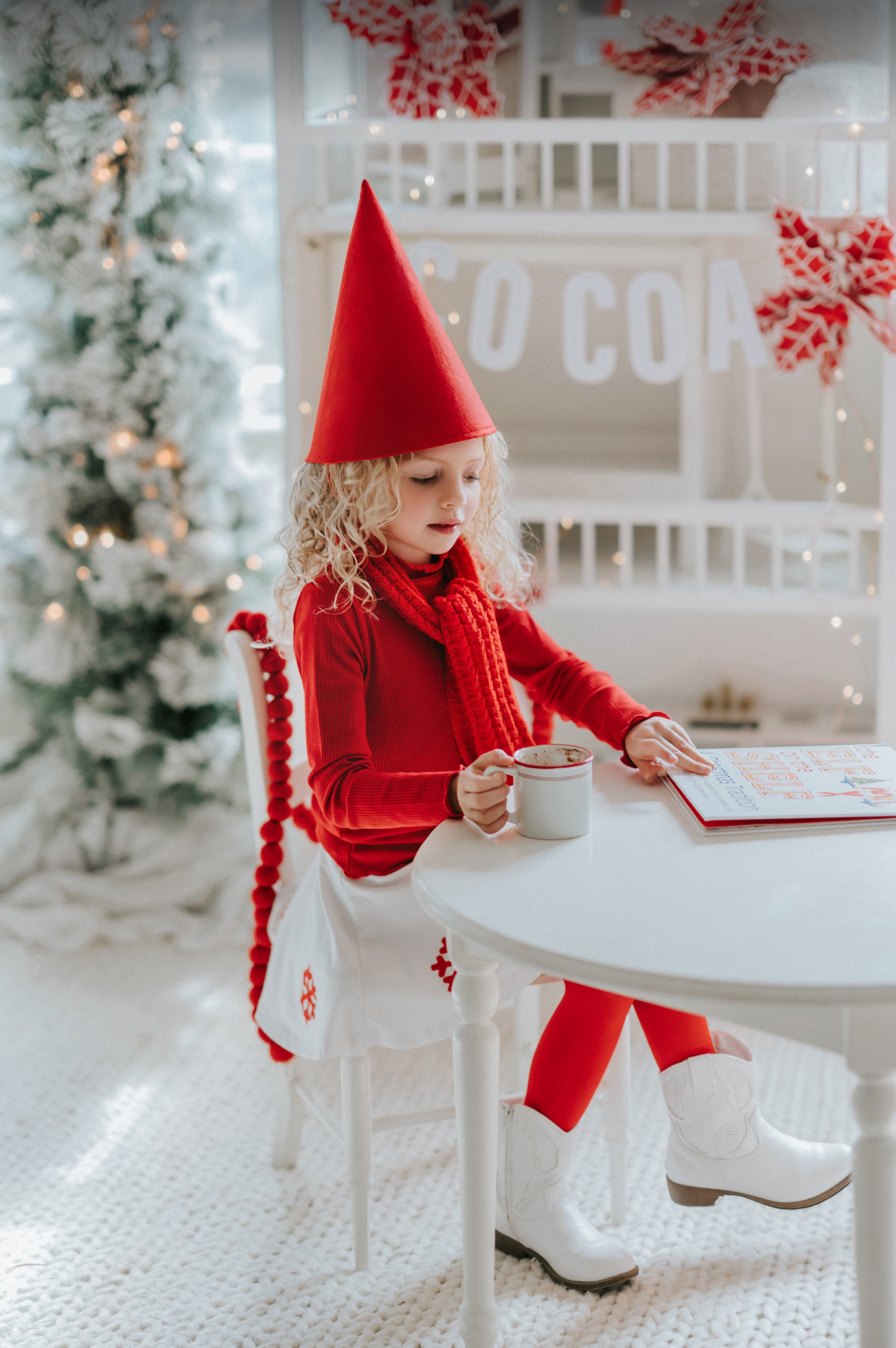 A young girl is wearing a red pointy gnome hat with a white round shape skirt. There are red snowflake applique around the bottom of the skirt.