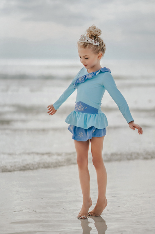 A young girl stands wearing a light blue 2 piece rash guard swim set.  The long sleeve top has an asymetric cornflower blue ruffle with silver snow flake accents. The skirted bottom is cornflower blue and has silver snowflake.