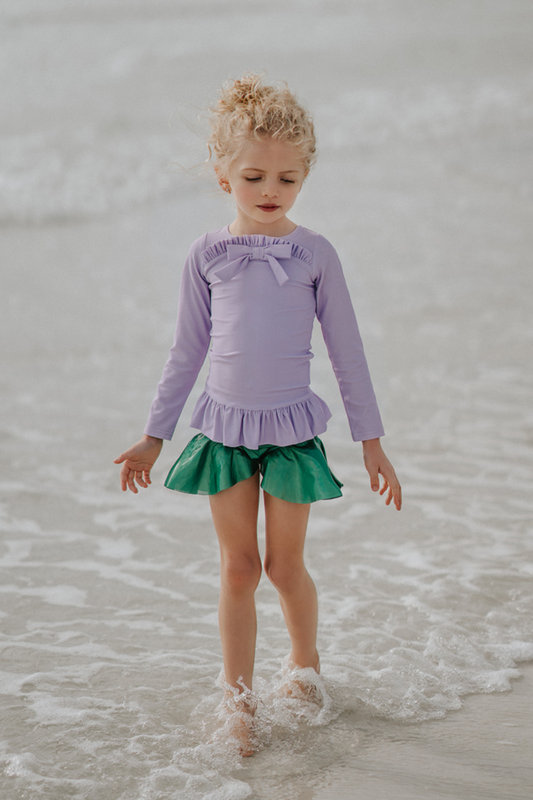 A young child walking on the shoreline at the beach is wearing a Mermaid Princess Rash Guard Swim Set with a long-sleeve purple top with a ruffle and  bow at the chest.  The modest bottom is metallic green and flares out at the sides to mimic a mermaid tail.  The swim suit offers SPF 50 for protection from the sun. 