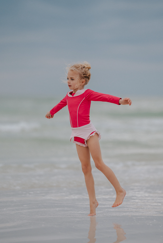 A young girl is wearing a pink 2 piece rashgaurd swim set.  The Sleeping Beauty swim set has a pink collar that resembles a ballgown.  The top has light pink piping and trim.  The modest bottoms have a pink overlay and ruffles on the legs. 