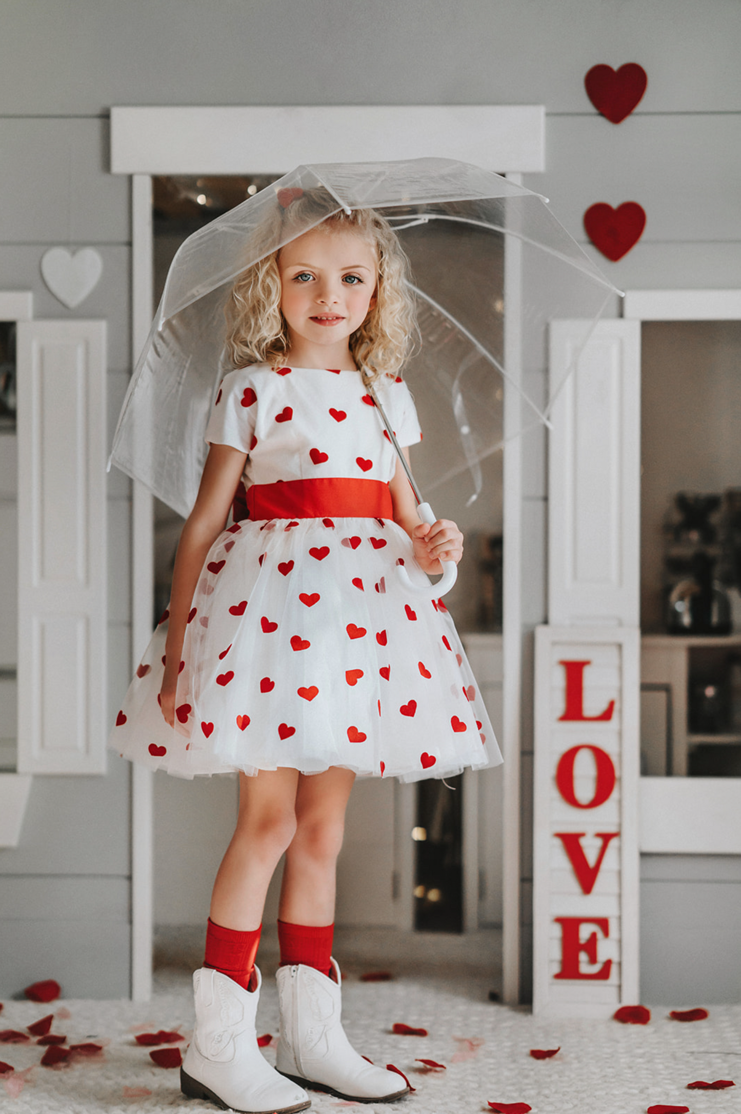 A young girl stands under a transparent umbrella. She wears the Sweet Little Hearts Frock, featuring red hearts and a red waist sash that ties in the back.  The short sleeve dress is white and has layers of tulle for a fuller skirt. 