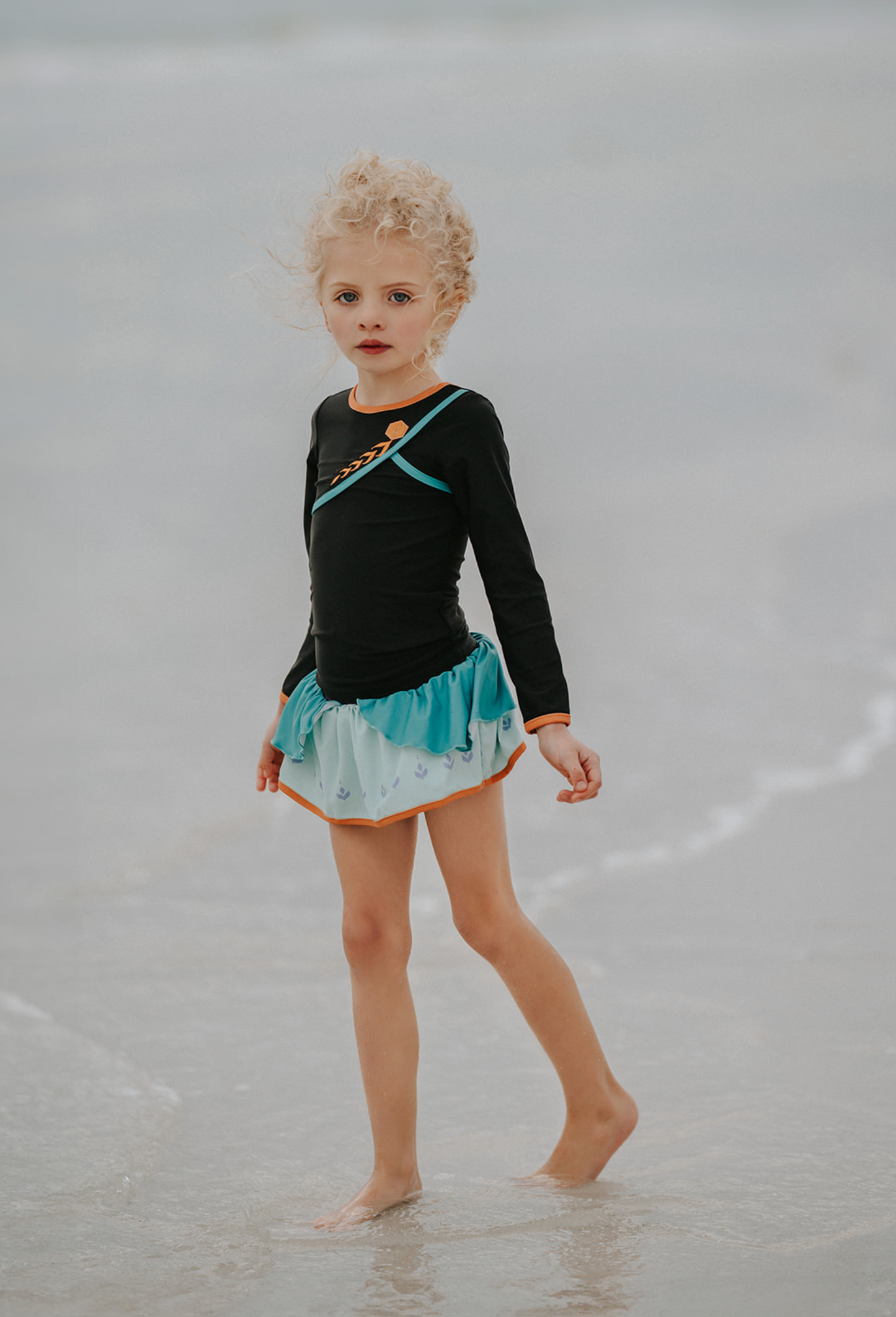 A young girl is wearing a 2 piece long sleeve rash guard swim set. The top is black with turquoise and goldenrod accents. The bottom has a turquoise ruffle over a mint teal skirt with goldenrod hem accent. 