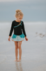 A young girl is wearing a 2 piece long sleeve rash guard swim set. The top is black with turquoise and goldenrod accents. The bottom has a turquoise ruffle over a mint teal skirt with goldenrod hem accent. 