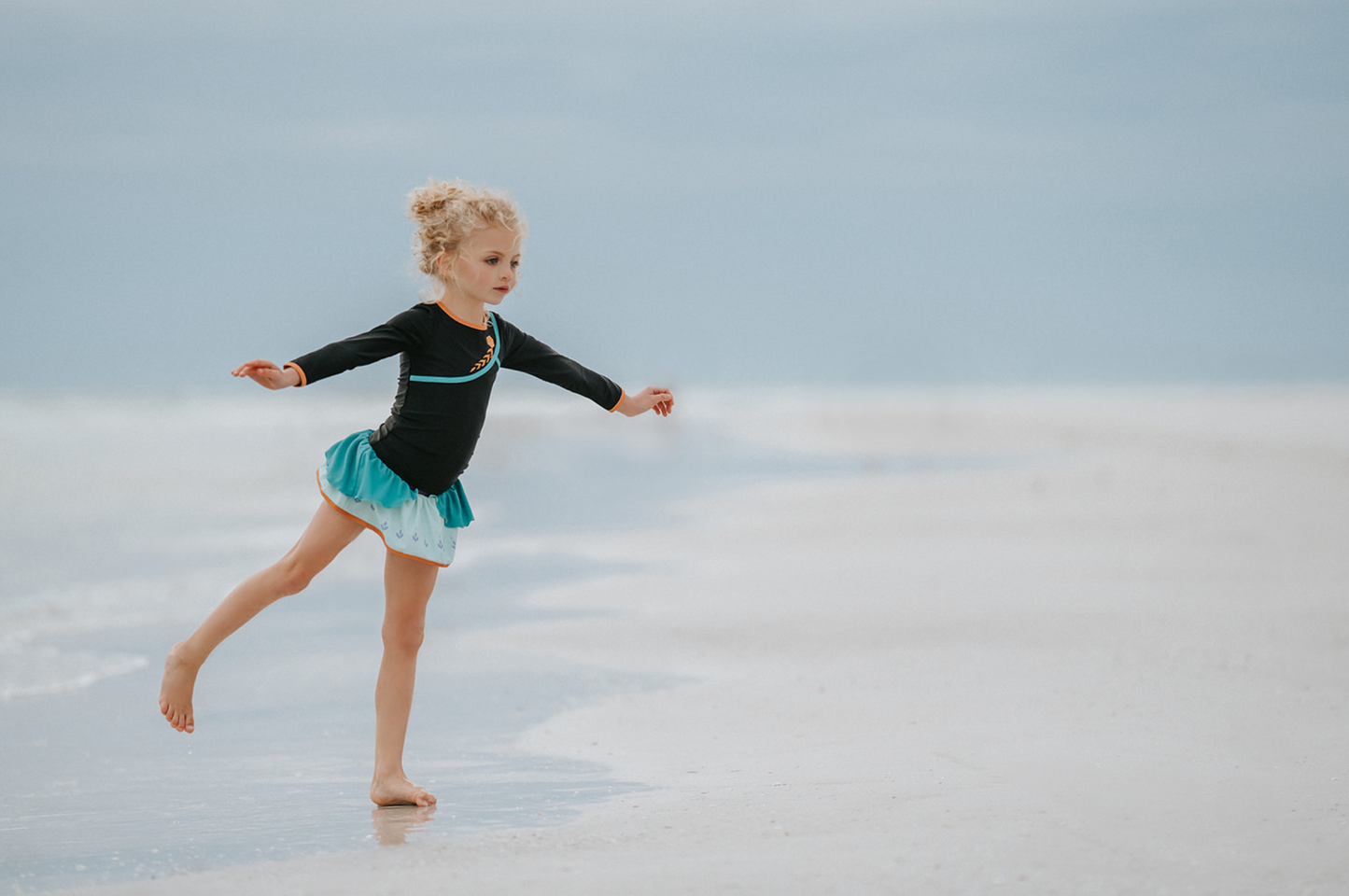 A young girl is wearing a 2 piece long sleeve rash guard swim set. The top is black with turquoise and goldenrod accents. The bottom has a turquoise ruffle over a mint teal skirt with goldenrod hem accent. 