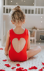 A young girl is wearing the Love Bug Leotard, a red gymnastics outfit with an elegant heart cutout back. 