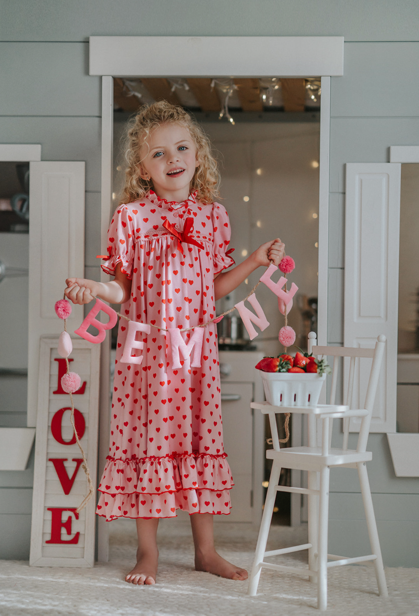 A young girl is wearing The Original Clara Nutcracker Gown in happy hearts print, featuring pink and red ruffles and a  red bow. The buttery soft fabric is cozy and sensory friendly. The pink gown has red hearts all over. The puff sleeves with ribbon detail and the ruffles at the hem add a traditional classic look. 